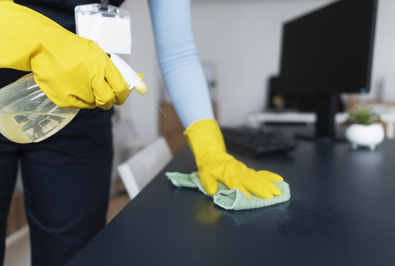 Main dans un gant bleu nettoyant une surface avec un chiffon gris, éponge jaune en arrière-plan.
