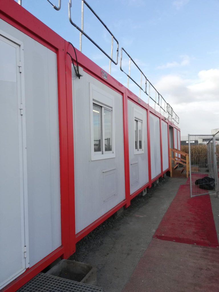 Bâtiment préfabriqué rouge et blanc, avec fenêtres, sur un chemin de sable.